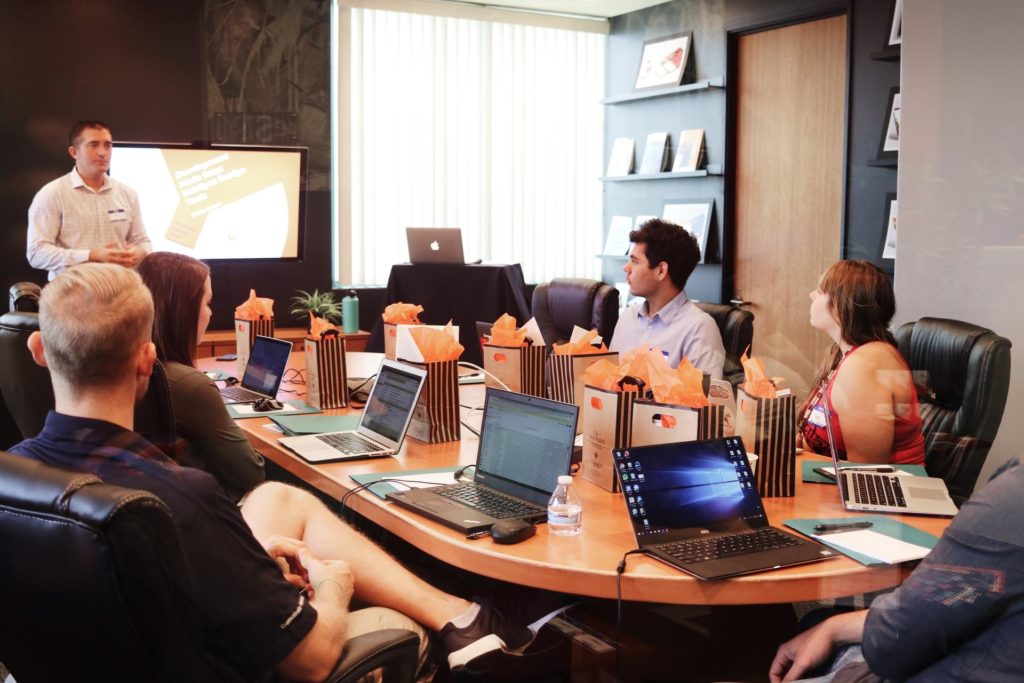 A corporate meeting room full of people listening to a presentation. There are laptops and gift bags scattered across the table.