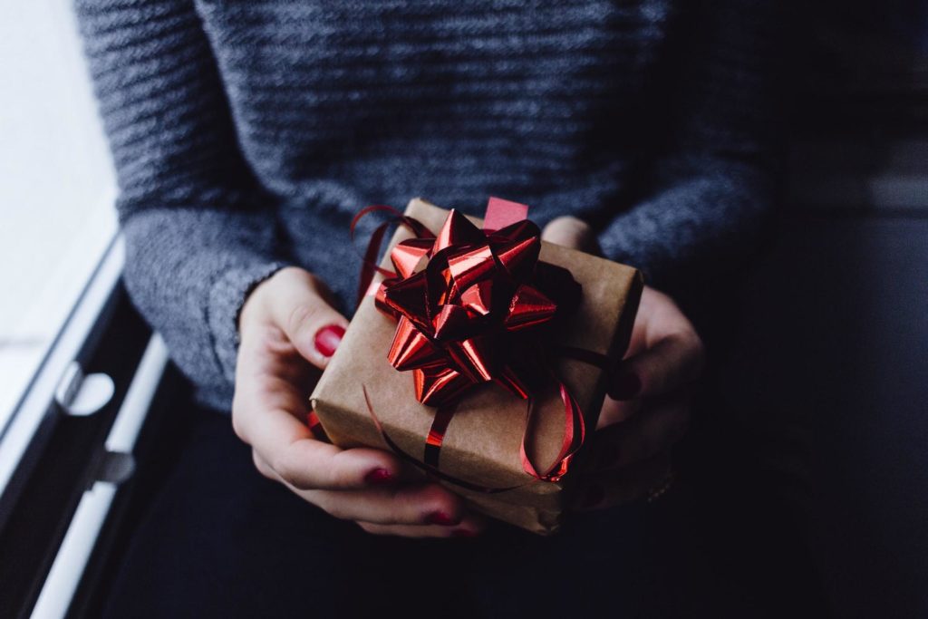 A woman in a dark sweater offers a present wrapped in brown paper with a red bow on it to the camera.